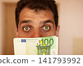 Close-up portrait of surprised young man with wide open eyes holding stack of one hundred euro banknotes near his face in blur. Concept of money, financial success, savings, wealth, income 141793992
