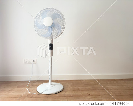 A white standing electric fan plugged into a wall socket on a round base. Home cooling appliance for summer days A white standing electric fan plugged into a wall socket on a round base. Home cooling appliance for summer days 141794004