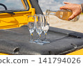 Close-up shot of man's hand pouring sparkling white wine or champagne from bottle into elegant glass flutes. Glasses stand on black textured hood of yellow off-road vehicle during an outdoor picnic  141794026