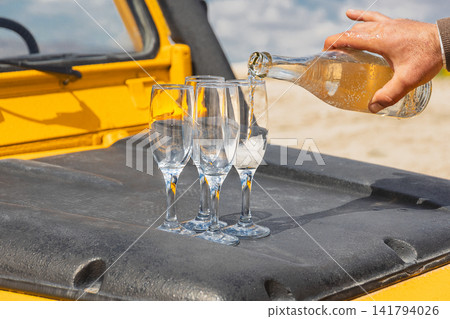 Close-up shot of man's hand pouring sparkling white wine or champagne from bottle into elegant glass flutes. Glasses stand on black textured hood of yellow off-road vehicle during an outdoor picnic Close-up shot of man's hand pouring sparkling white wine or champagne from bottle into elegant glass flutes. Glasses stand on black textured hood of yellow off-road vehicle during an outdoor picnic 141794026