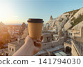 First person view of hand holding craft paper takeaway coffee cup against background of unique cave houses and volcanic rocks in Cappadocia, Turkey at sunrise 141794030