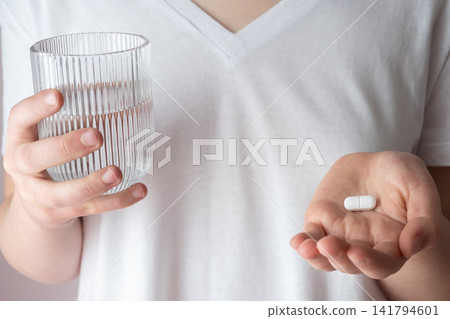 Girl in white clothes take the pill and glass with water in the hand. Healthcare from pharmacy. Kid sit at home because of illness. Girl in white clothes take the pill and glass with water in the hand. Healthcare from pharmacy. Kid sit at home because of illness. 141794601