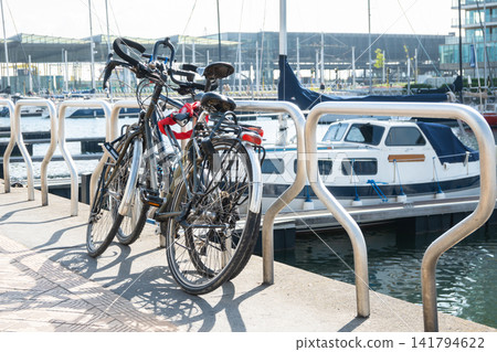 Bicycles parking on the street near the sea port with boats and yachts. Blue sea, houses and summer sky. Bike for sport or travel in Ostende, Belgium. Bicycles parking on the street near the sea port with boats and yachts. Blue sea, houses and summer sky. Bike for sport or travel in Ostende, Belgium. 141794622