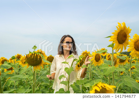 Young woman in sunglasses holding large sunflower amid bright summer field under clear blue sky, relaxed pose with warm natural light and vibrant yellow blooms surrounding her Young woman in sunglasses holding large sunflower amid bright summer field under clear blue sky, relaxed pose with warm natural light and vibrant yellow blooms surrounding her 141794677