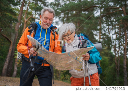 Hiking tourism. Senior couple man woman looking track in paper map enjoying hiking in forest. Happy old people backpackers hikers enjoy walking hike trekking tourism active vacation beauty of nature Hiking tourism. Senior couple man woman looking track in paper map enjoying hiking in forest. Happy old people backpackers hikers enjoy walking hike trekking tourism active vacation beauty of nature 141794913