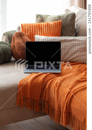 Laptop on couch with textured pillows and orange throw blanket cozy home comfort Laptop on couch with textured pillows and orange throw blanket cozy home comfort 141795806