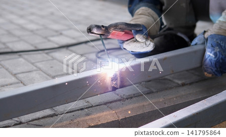 welder wearing protective gear welding metal beam in outdoor construction site with concentrated industrious mood and vivid colors of gray, blue, and red, close-up of welding process, ideal. welder wearing protective gear welding metal beam in outdoor construction site with concentrated industrious mood and vivid colors of gray, blue, and red, close-up of welding process, ideal. 141795864