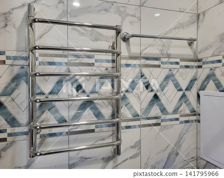 In the bathroom, a metal towel warmer hangs on the wall made of gray tiles. In the bathroom, a metal towel warmer hangs on the wall made of gray tiles. 141795966