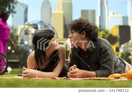 Young couple laying face to face and smiling at each other on picnic blanket, smiling during intimate conversation with food and blurred city skyline. Romantic picnic idea outdoors. 141796232