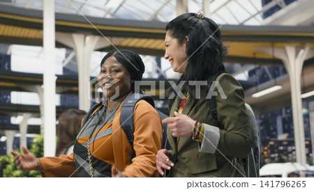 Happy women walking together through modern bus terminal while chatting. Cheerful female tourists carrying backpacks in shuttle station enjoying friendly conversation before departure Happy women walking together through modern bus terminal while chatting. Cheerful female tourists carrying backpacks in shuttle station enjoying friendly conversation before departure 141796265