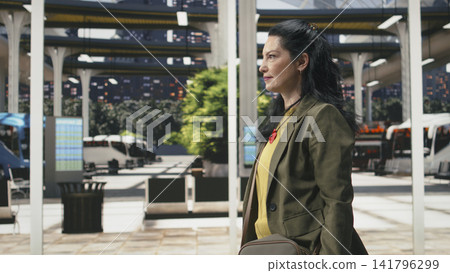Traveler walking in bus station taking ticket out of bag to check departure time. Female passenger looking at wrist watch to make sure she is not late Traveler walking in bus station taking ticket out of bag to check departure time. Female passenger looking at wrist watch to make sure she is not late 141796299