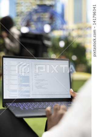 Close up of person working with programming interface and source code on laptop, representing software development and flexible IT work in city park. Over-the-shoulder view. Close up of person working with programming interface and source code on laptop, representing software development and flexible IT work in city park. Over-the-shoulder view. 141796341