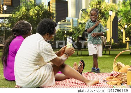 African dad holding mobile phone to take photos of child posing in city park, capturing family memory beside picnic blanket downtown. Proud father taking pictures of kid. African dad holding mobile phone to take photos of child posing in city park, capturing family memory beside picnic blanket downtown. Proud father taking pictures of kid. 141796589
