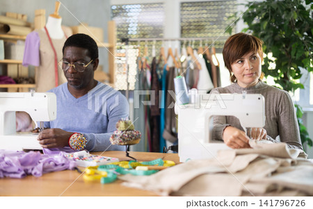 Woman tailor and African male companion sew clothes in sewing machine Woman tailor and African male companion sew clothes in sewing machine 141796726