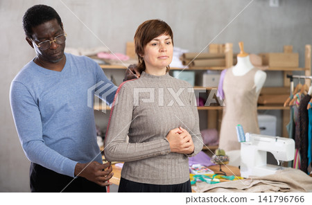 Male tailor using measuring tape to measure clothes on woman in sewing studio Male tailor using measuring tape to measure clothes on woman in sewing studio 141796766