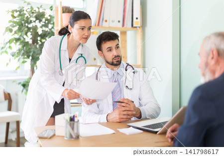Senior man client of medical clinic sits opposite male and female doctor and communicates with specialists Senior man client of medical clinic sits opposite male and female doctor and communicates with specialists 141798617