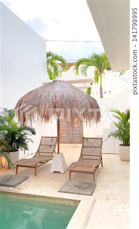 Tropical courtyard retreat with thatched palapa umbrella, twin woven lounge chairs, turquoise pool, white architecture, palm trees, pale stone tiles and relaxed resort setting Tropical courtyard retreat with thatched palapa umbrella, twin woven lounge chairs, turquoise pool, white architecture, palm trees, pale stone tiles and relaxed resort setting 141798995