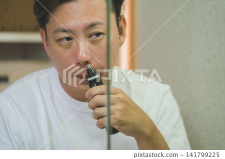A man in his 40s using a nose trimmer over the edge of a bathroom mirror. A man in his 40s using a nose trimmer over the edge of a bathroom mirror. 141799225