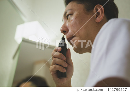 一位四十多歲的男子在浴室鏡子前使用鼻毛修剪器(側面視圖)。 一位四十多歲的男子在浴室鏡子前使用鼻毛修剪器(側面視圖)。 141799227