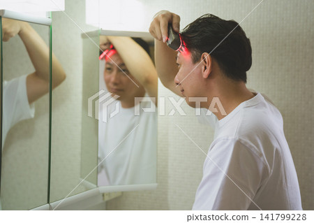 一名 40 歲的男子在浴室鏡子前使用 LED 頭皮按摩刷進行頭皮護理。 一名 40 歲的男子在浴室鏡子前使用 LED 頭皮按摩刷進行頭皮護理。 141799228