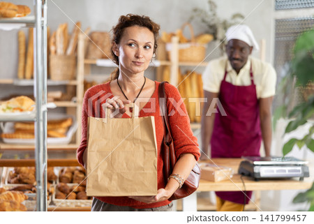 Woman buyer with pastry and baguettes in paper package stands near glass showcase in bakery Woman buyer with pastry and baguettes in paper package stands near glass showcase in bakery 141799457