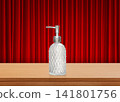 A clear glass perfume bottle sits on a wooden table, with a red curtain as a backdrop. 141801756