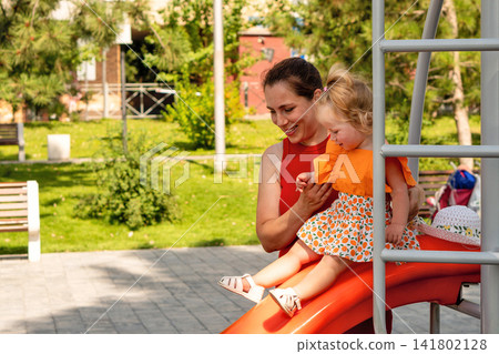 Smiling Mother Supporting Toddler Daughter on Orange Slide Smiling Mother Supporting Toddler Daughter on Orange Slide 141802128