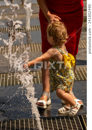 Toddler Girl Reaching Toward Fountain With Mother Nearby Toddler Girl Reaching Toward Fountain With Mother Nearby 141802490