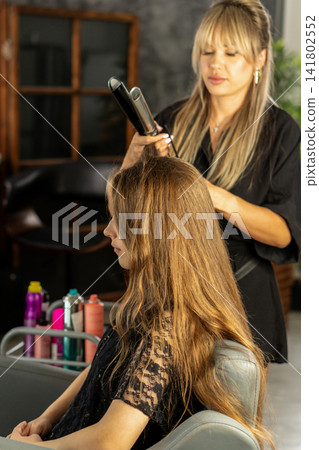 Hairstylist Straightening Hair Of Teen Client In Beauty Salon Hairstylist Straightening Hair Of Teen Client In Beauty Salon 141802552