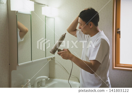 A man in his 40s using a hairdryer to style his hair in front of the bathroom mirror. A man in his 40s using a hairdryer to style his hair in front of the bathroom mirror. 141803122