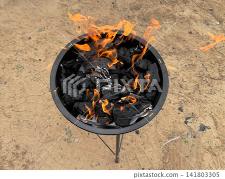 Charcoal burned in a portable grill on sandy ground for use in outdoor barbecues or camp cooking. Charcoal burned in a portable grill on sandy ground for use in outdoor barbecues or camp cooking. 141803305