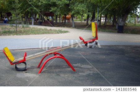 A vibrant red and yellow seesaw sits on a dark playground surface. In the background of this park, green trees, park benches. A vibrant red and yellow seesaw sits on a dark playground surface. In the background of this park, green trees, park benches. 141803546