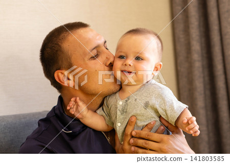 Father Kissing Smiling Baby Son On Cheek Indoors Father Kissing Smiling Baby Son On Cheek Indoors 141803585