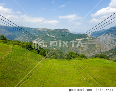 Cable car cables cross lush green slopes toward rocky mountain ridges under clea Cable car cables cross lush green slopes toward rocky mountain ridges under clea 141803654