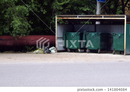 Three large green wheeled dumpsters sit under a metal-framed shelter on a roadside. Loose trash is next to them on the dirt and asphalt. Background against dense green foliage. Three large green wheeled dumpsters sit under a metal-framed shelter on a roadside. Loose trash is next to them on the dirt and asphalt. Background against dense green foliage. 141804084