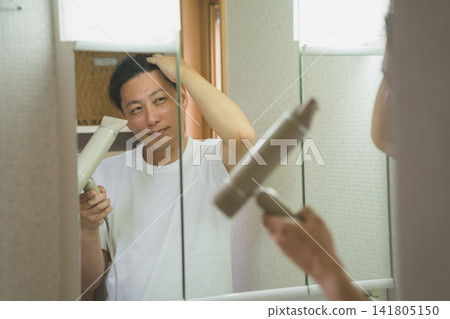 A man in his 40s drying his hair with a hairdryer in front of the bathroom mirror. A man in his 40s drying his hair with a hairdryer in front of the bathroom mirror. 141805150