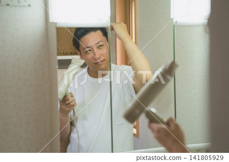 浴室鏡子裡映出一位面帶微笑的四十多歲男子,他正在用吹風機吹乾頭髮。 浴室鏡子裡映出一位面帶微笑的四十多歲男子,他正在用吹風機吹乾頭髮。 141805929