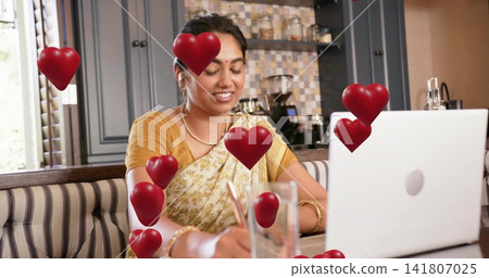 Writing woman in saree using laptop and pen at kitchen nook, with red heart overlays 141807025