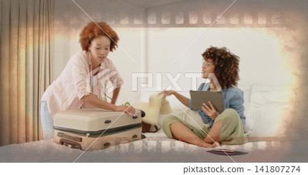 Packing two women in denim shirt closing beige suitcase on bed in bright bedroom, with tablet 141807274