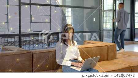Typing woman in white blouse and jeans on sofa in coworking lounge using silver laptop, headset Typing woman in white blouse and jeans on sofa in coworking lounge using silver laptop, headset 141807478