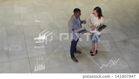 Discussing, two staff in business attire reviewing laptop, documents and projected charts in atrium Discussing, two staff in business attire reviewing laptop, documents and projected charts in atrium 141807947