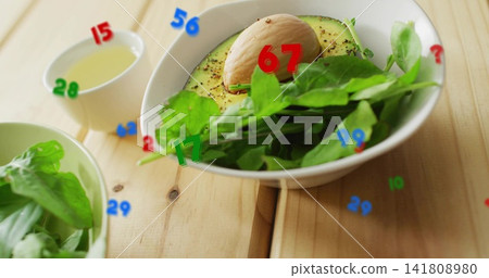 Displaying white bowl holding halved avocado with pit, greens on wood table, with numeric overlays Displaying white bowl holding halved avocado with pit, greens on wood table, with numeric overlays 141808980