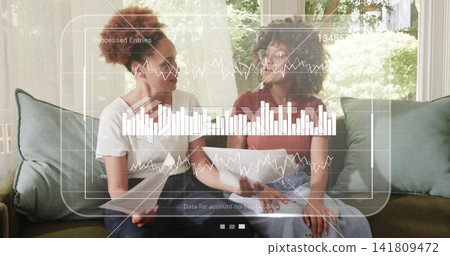Reviewing docs, two women sitting on green sofa at home wearing short sleeves, viewing data overlay Reviewing docs, two women sitting on green sofa at home wearing short sleeves, viewing data overlay 141809472