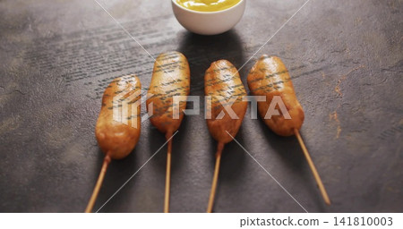 Displaying four battered corn dogs on wooden skewers resting on dark slate tabletop, with mustard Displaying four battered corn dogs on wooden skewers resting on dark slate tabletop, with mustard 141810003