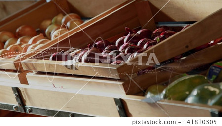 Displaying wooden crate holding glossy red onions on market shelf, with shallots, green fruits, tag 141810305