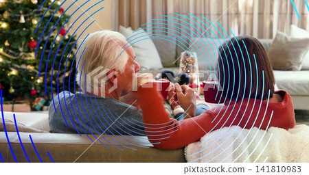Sipping senior couple wearing sweaters, sitting on sofa at home with blue overlay, holding red mugs Sipping senior couple wearing sweaters, sitting on sofa at home with blue overlay, holding red mugs 141810983