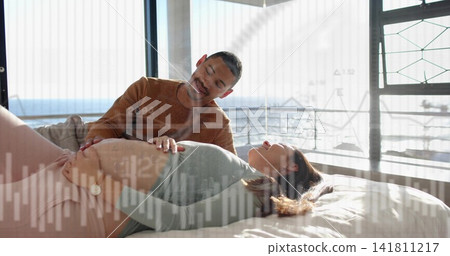 Smiling partner leaning over pregnant woman in green top, placing hand on belly on bed, overlays 141811217