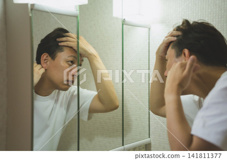 一名40歲的男子因頭髮稀疏而感到擔憂,他在浴室洗手台旁接受了雄性禿(AGA)檢查。 一名40歲的男子因頭髮稀疏而感到擔憂,他在浴室洗手台旁接受了雄性禿(AGA)檢查。 141811377