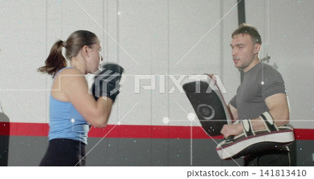 Throwing punches female trainee in blue tank, black gloves, male coach holding Thai pads in gym Throwing punches female trainee in blue tank, black gloves, male coach holding Thai pads in gym 141813410