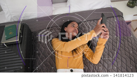 Lying woman in mustard top light pants holding smartphone on bed, with suitcase, passport and HUD Lying woman in mustard top light pants holding smartphone on bed, with suitcase, passport and HUD 141814123